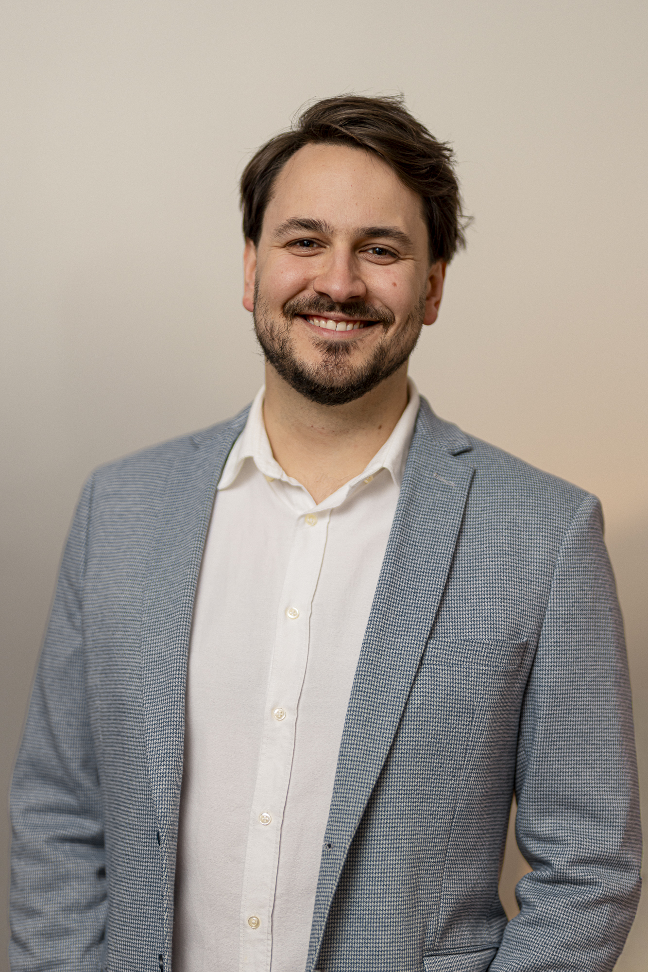Smiling man with beard wearing a light blue checked blazer and white shirt against a plain background.