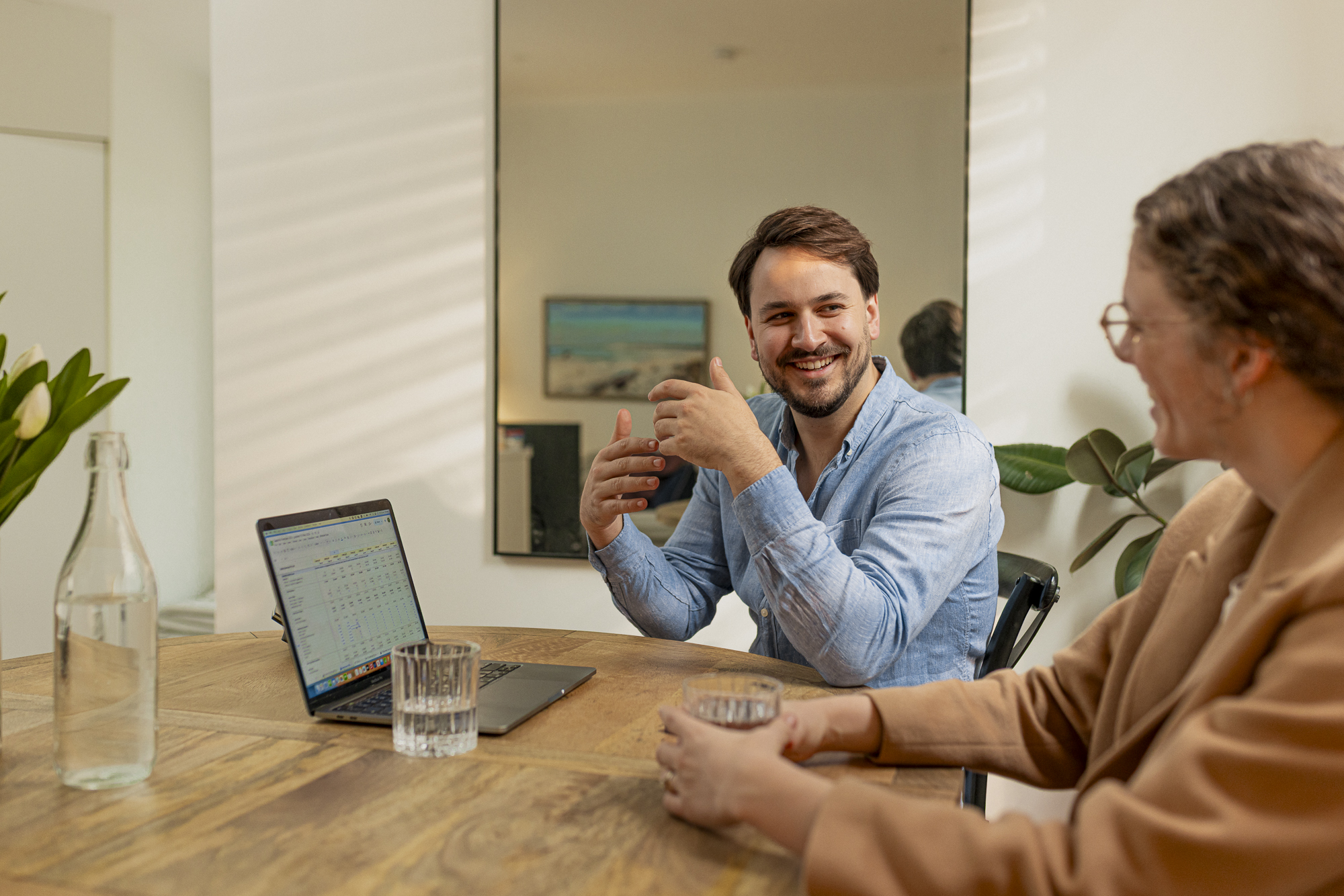 Two colleagues smiling and talking at a round table with a laptop and glasses of water.