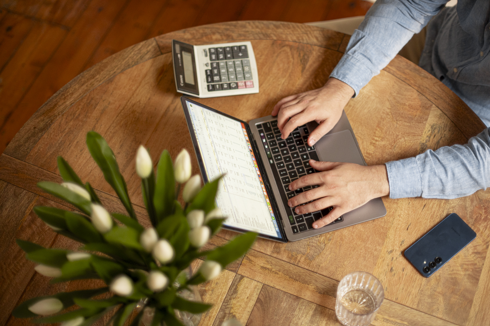 Person typing on laptop at wooden table with calculator, smartphone, glass of water, and blurred white flowers.