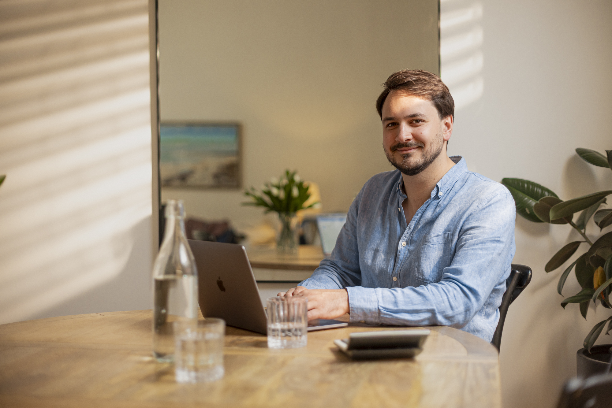 Man in blue shirt sitting at wooden table with laptop, glass bottle, and glasses, smiling at camera.