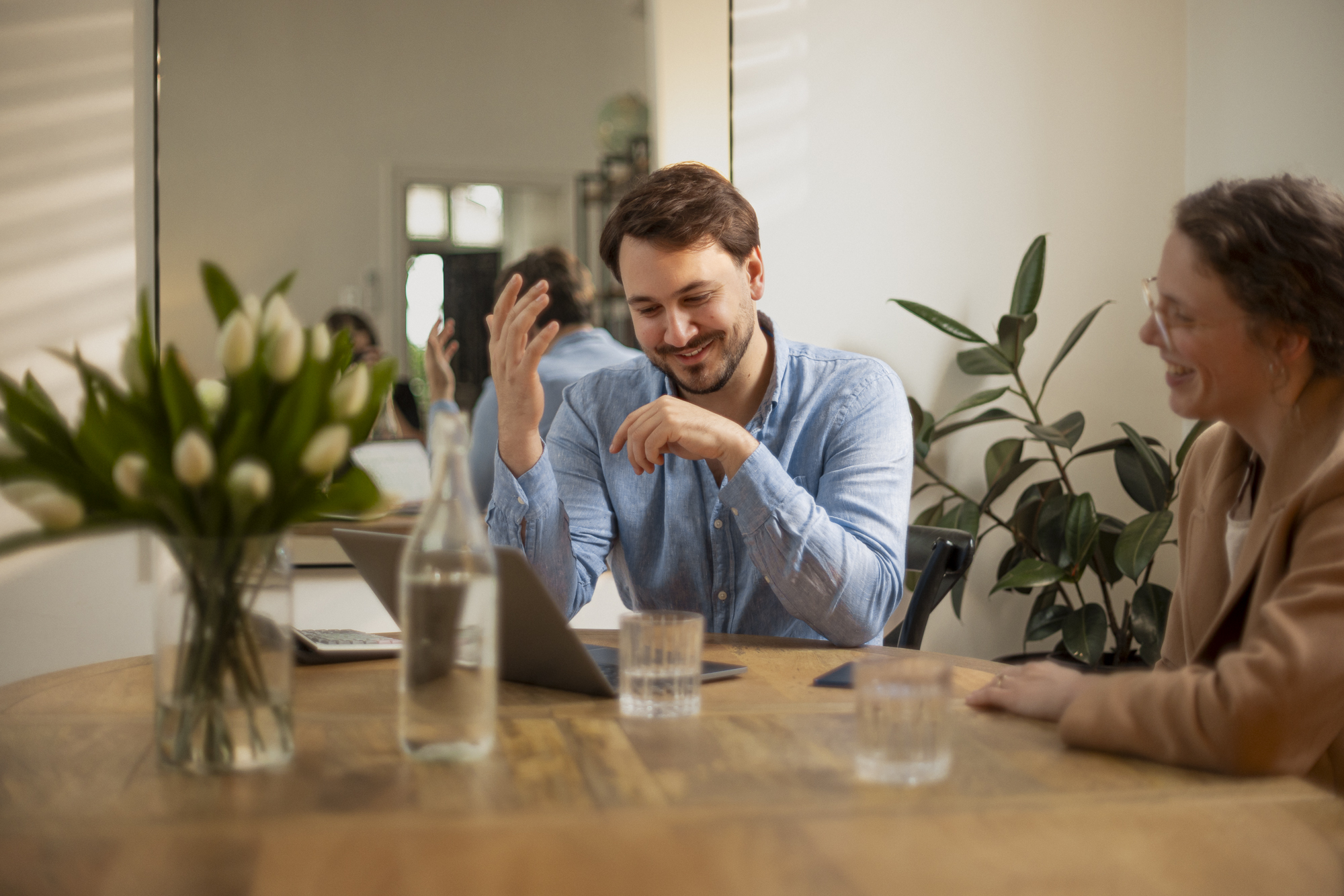Smiling man and woman sitting at a table with laptop, flowers, and water glasses in a light room.