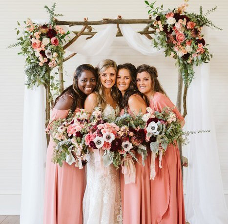 Women of the bridal party in front of arch wearing coral, holding bouquets. 