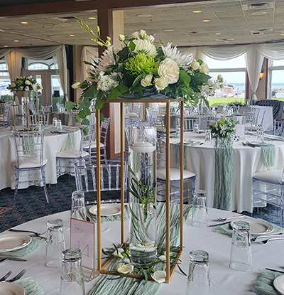Elegant wedding reception table centerpiece with white flowers and greenery on a gold stand, surrounded by glassware and white linens.