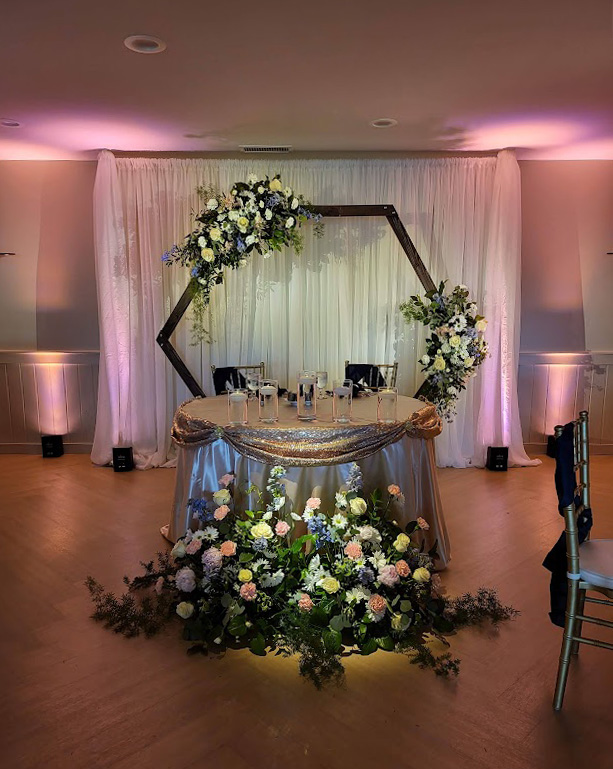 White backdrop with arch and floral decoration behind a head table. The head table surrounded by flowers. 