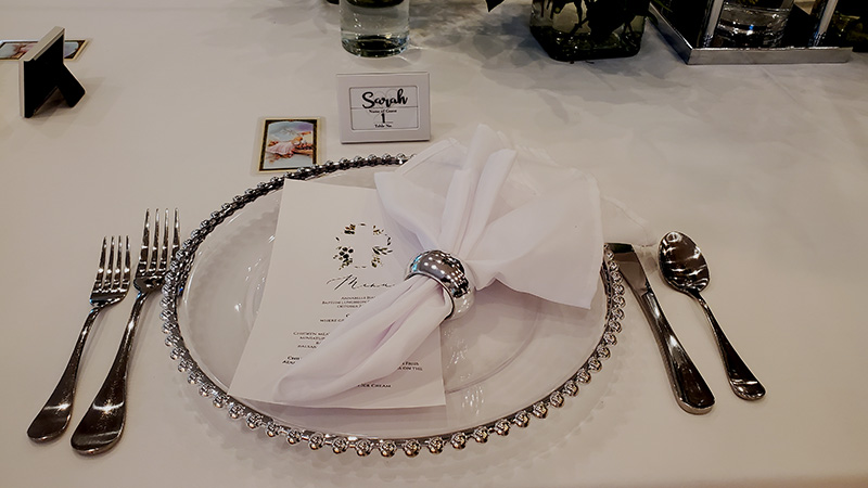 A clear charger with a silver dotted rim, silver flatware, a white napkin with a silver napkin ring, and a name card doubling as a menu. 