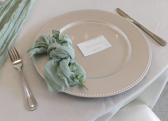 Silver charger, sage cheese cloth napkin, a name card, and silver flatware. 
