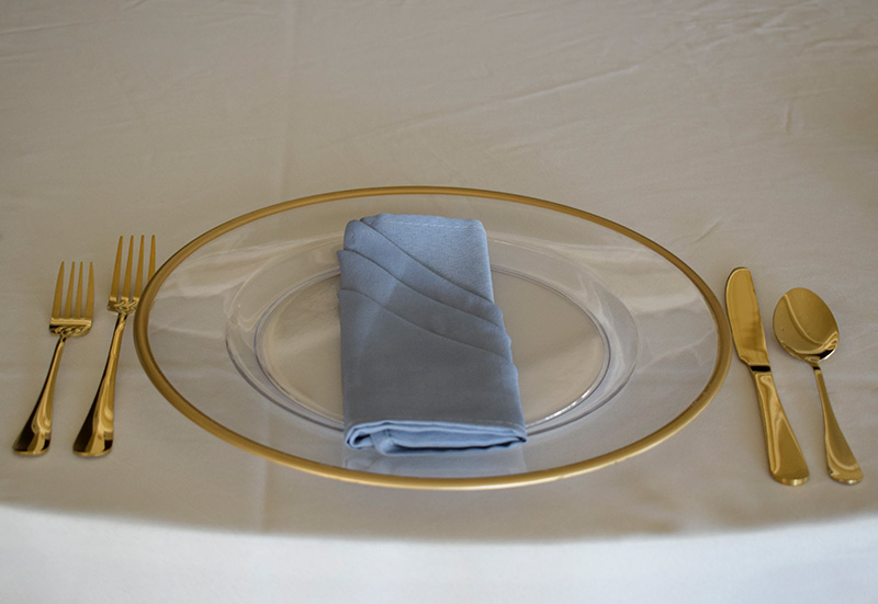 A clear charger with a gold rim, gold flatware, and a baby blue napkin. 