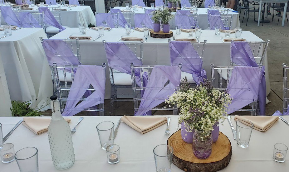 Centerpieces on wooden slabs made from lilac mason jars filled with Baby's Breath (flower name). Lilac chair sashes and Champaign colored napkins.  