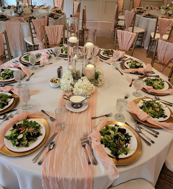 Salads on white plates on gold chargers with silver flatware and cylinder vases as centerpieces with rose cheesecloth runners and napkins. 