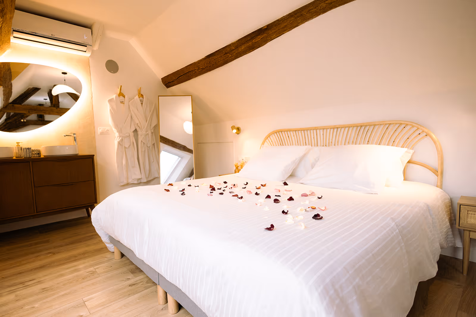 Chambre claire avec grand lit, têtes de lit en rotin, pétales sur la couette blanche, peignoirs et miroir.