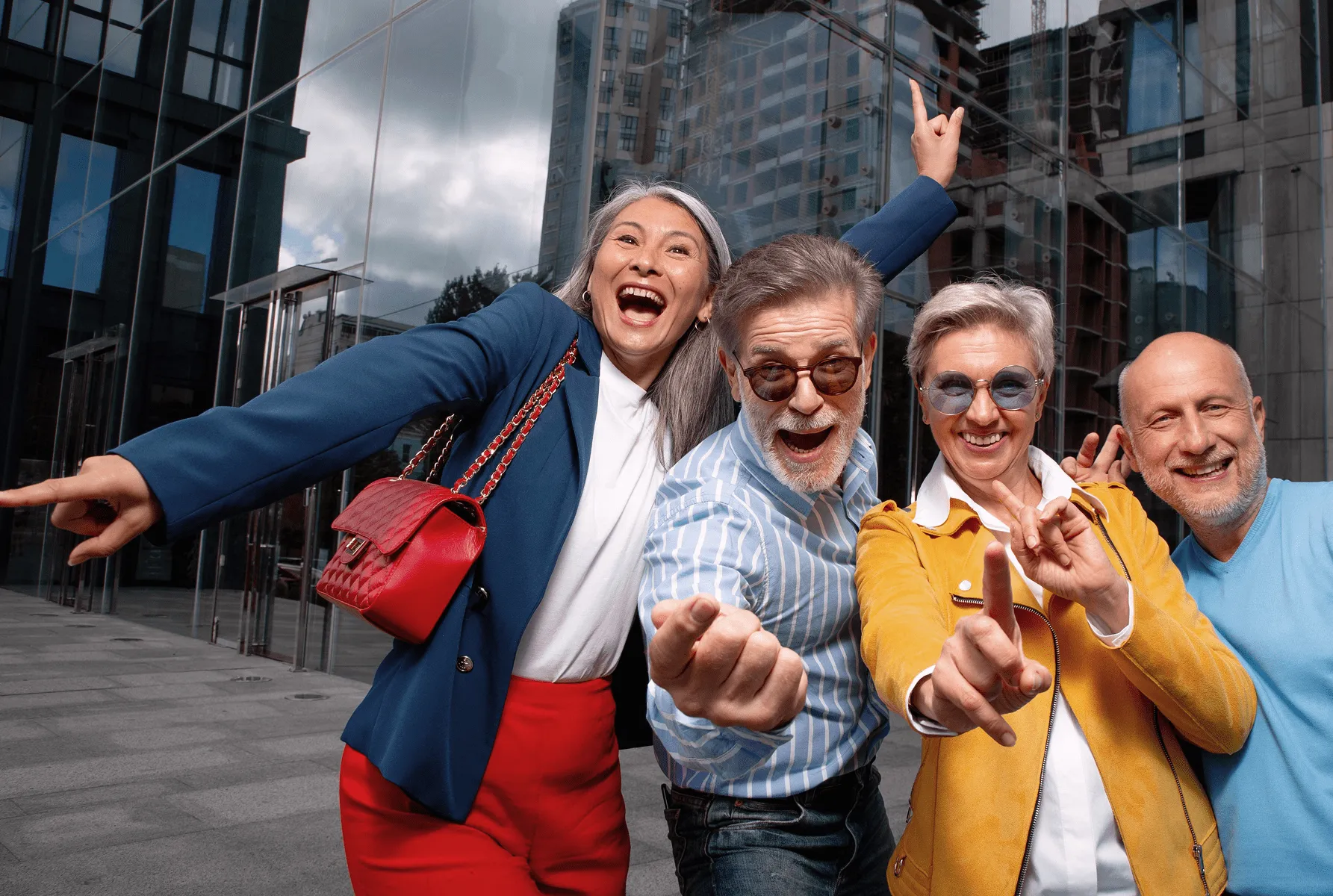 Četvorka srećnih starijih osoba sa naočarima pokazuje prste i osmehavaju se ispred staklene poslovne zgrade.
