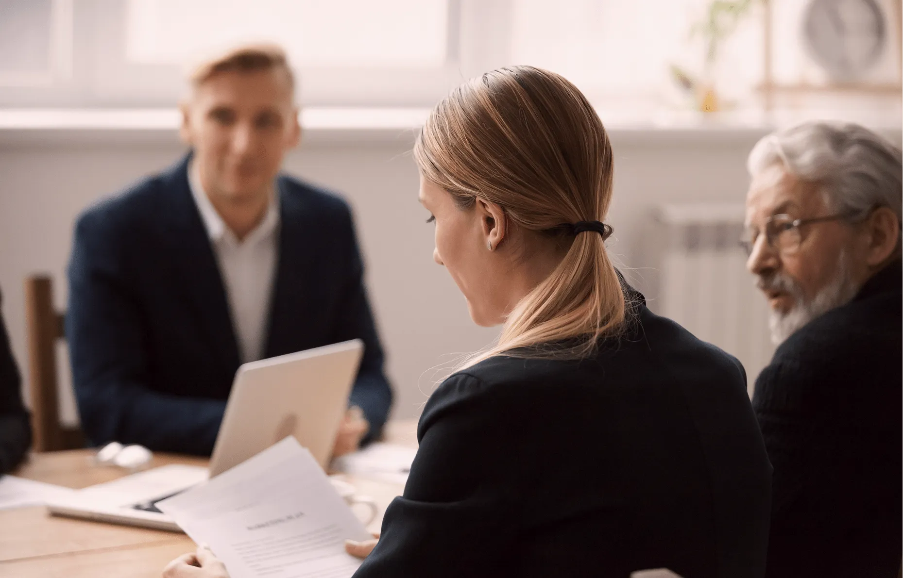 Grupa ljudi na poslovnom sastanku u kancelariji, žena drži dokument, muškarci razgovaraju.