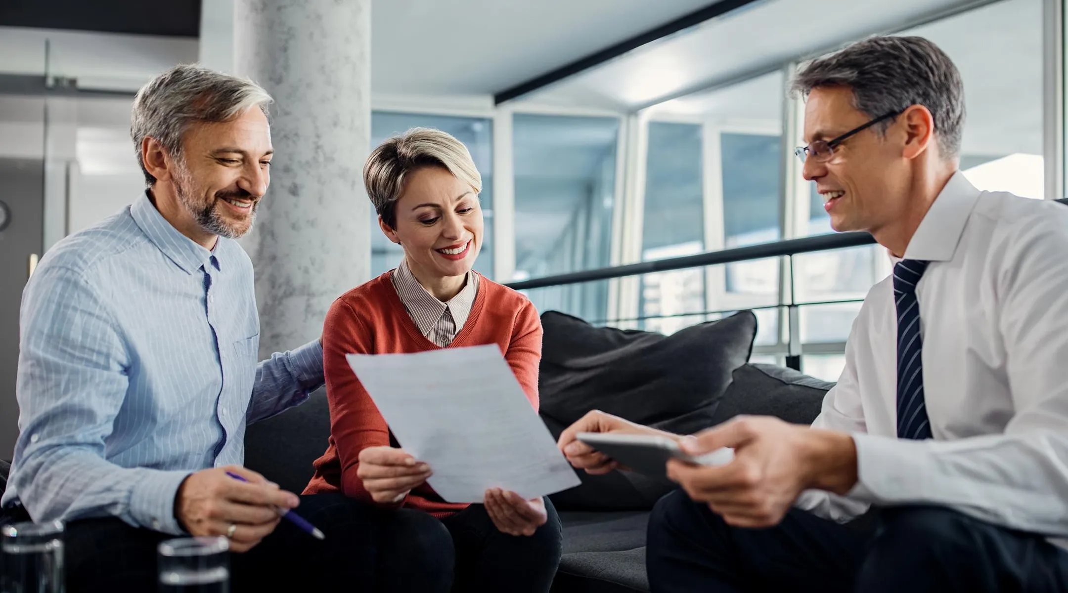 Dva muškarca i žena sede na kauču u modernom kancelarijskom prostoru dok pregledaju i diskutuju o dokumentu.