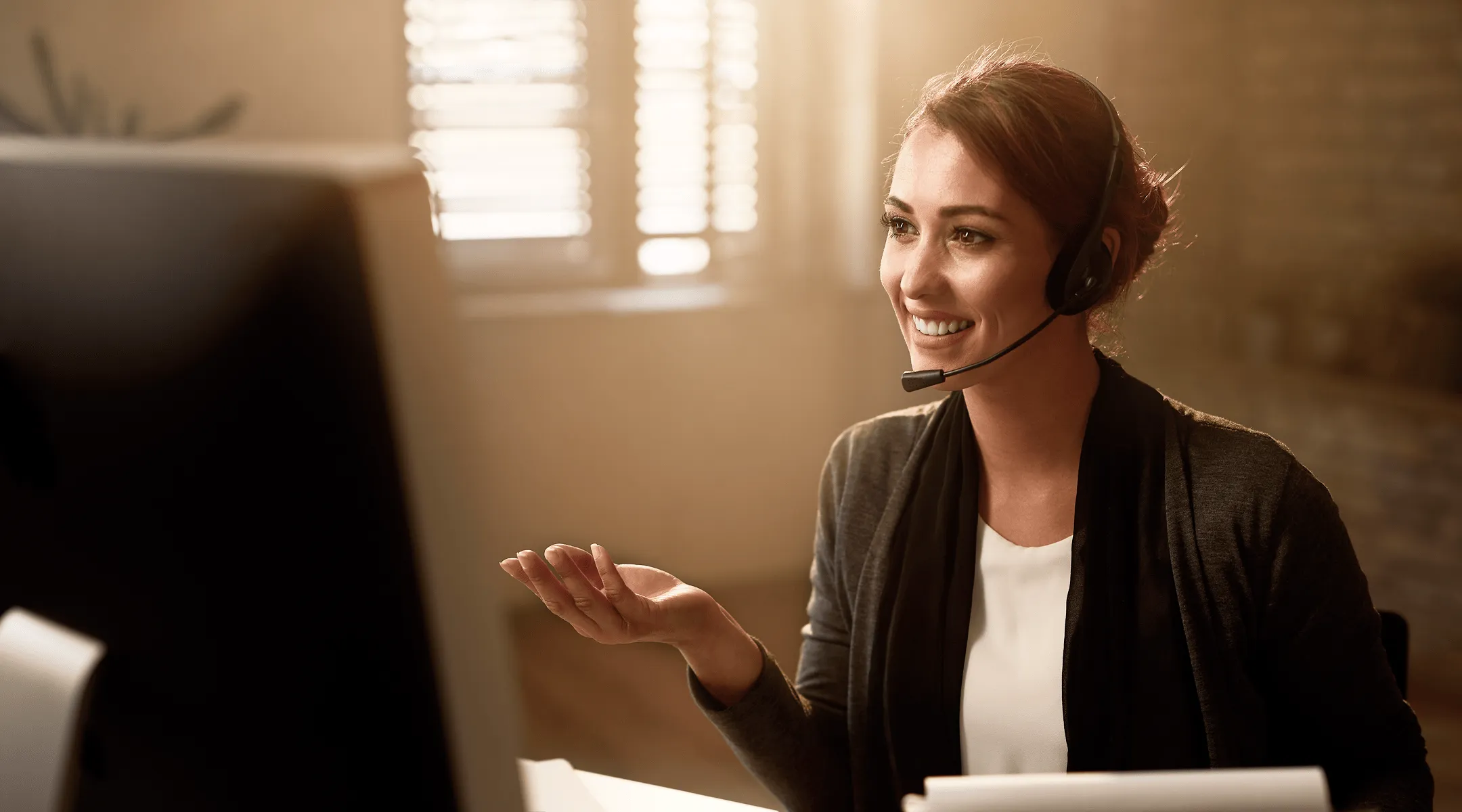 Смејућа се жена са слушалицама разговара преко рачунара у осветљеном канцеларијском простору.