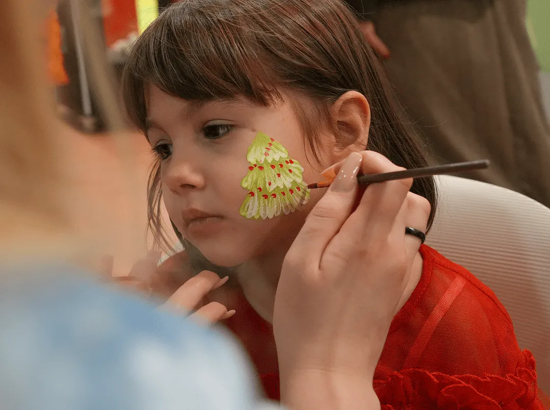 Девојчица обучена у црвено док јој лице фарбају сликарским кистом у дечији мотив, божићну јелку.