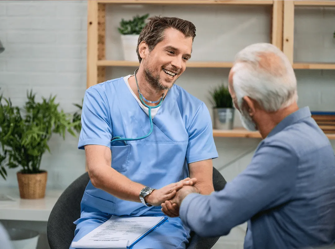 Medic u plavom uniformi nasmejan drži ruke starijeg muškarca tokom razgovora u kancelariji.