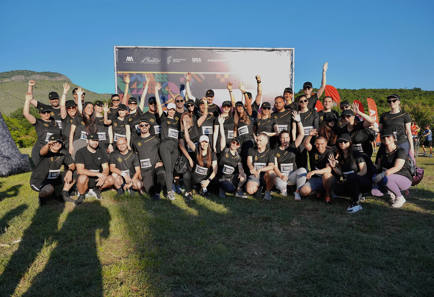 Велика група људи обучених у црне спортске мајице са натписом, окупљена напољу на зеленој ливади, весело поздравља и слика се испред платна са логотипима на ведром небу.