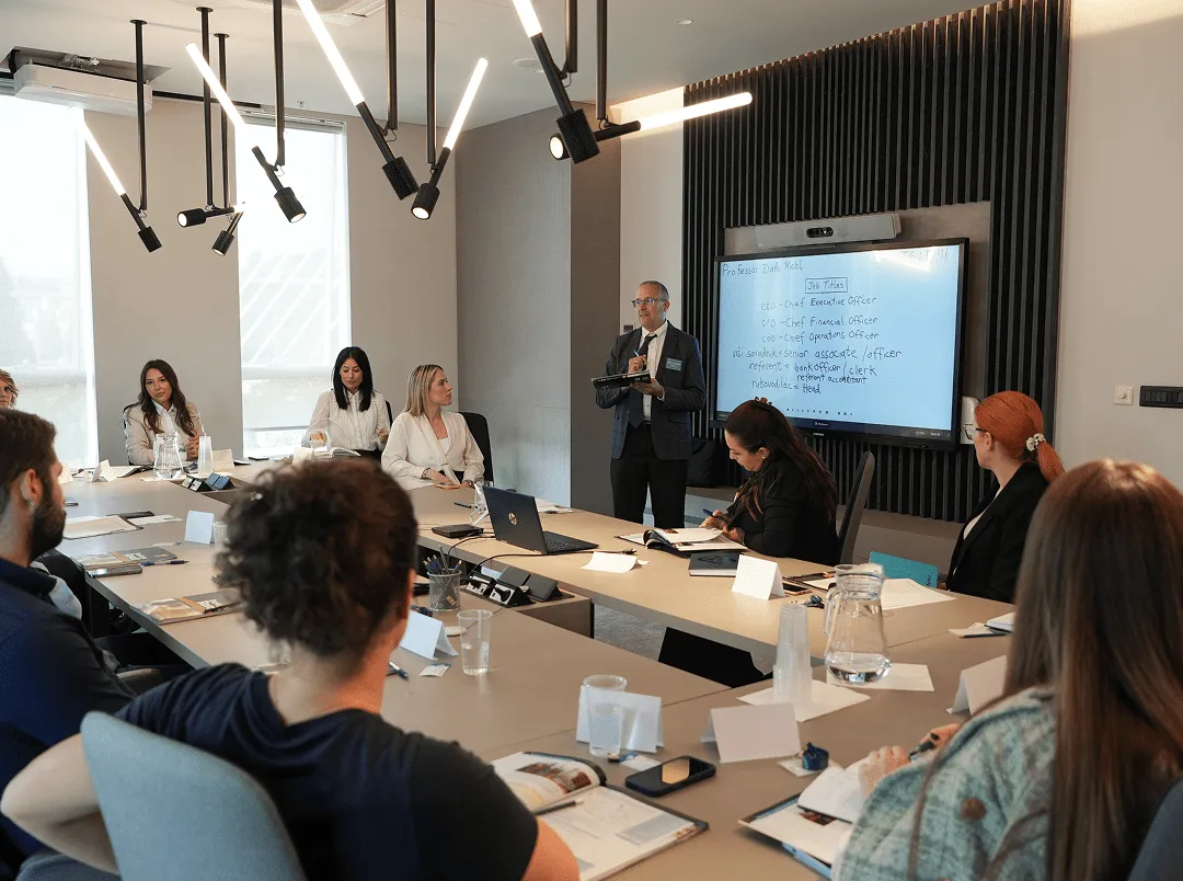 Muškarac u odelu drži prezentaciju pred grupom ljudi u modernoj konferencijskoj sali.