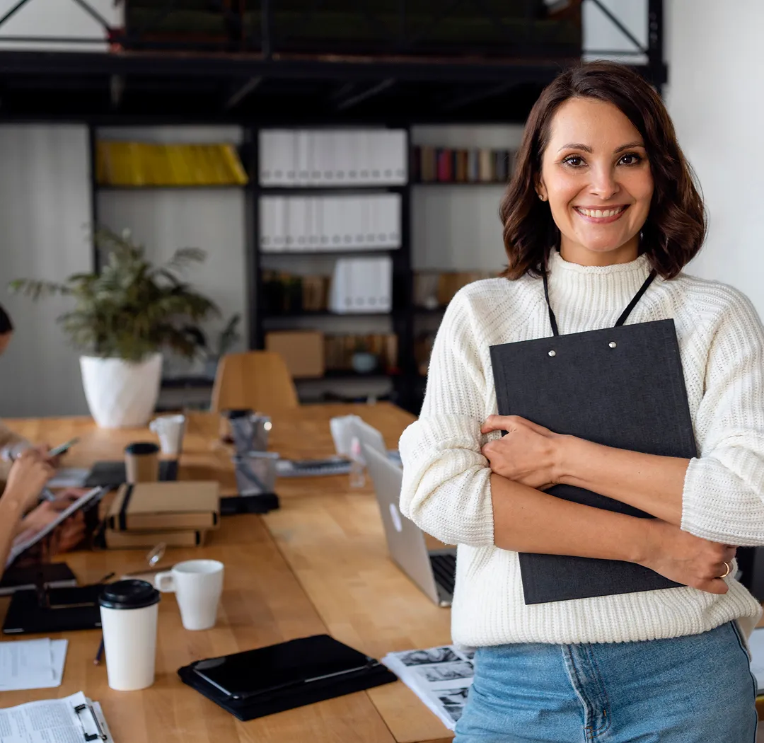 Žena u belom džemperu drži crnu fasciklu u kancelariji sa stolom punim dokumenata i laptopom.