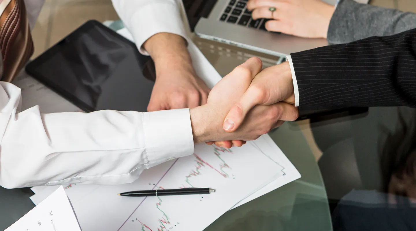 Dva muškarca se rukuju preko staklenog stola sa poslovnim dokumentima, olovkom, tabletom i laptopom.