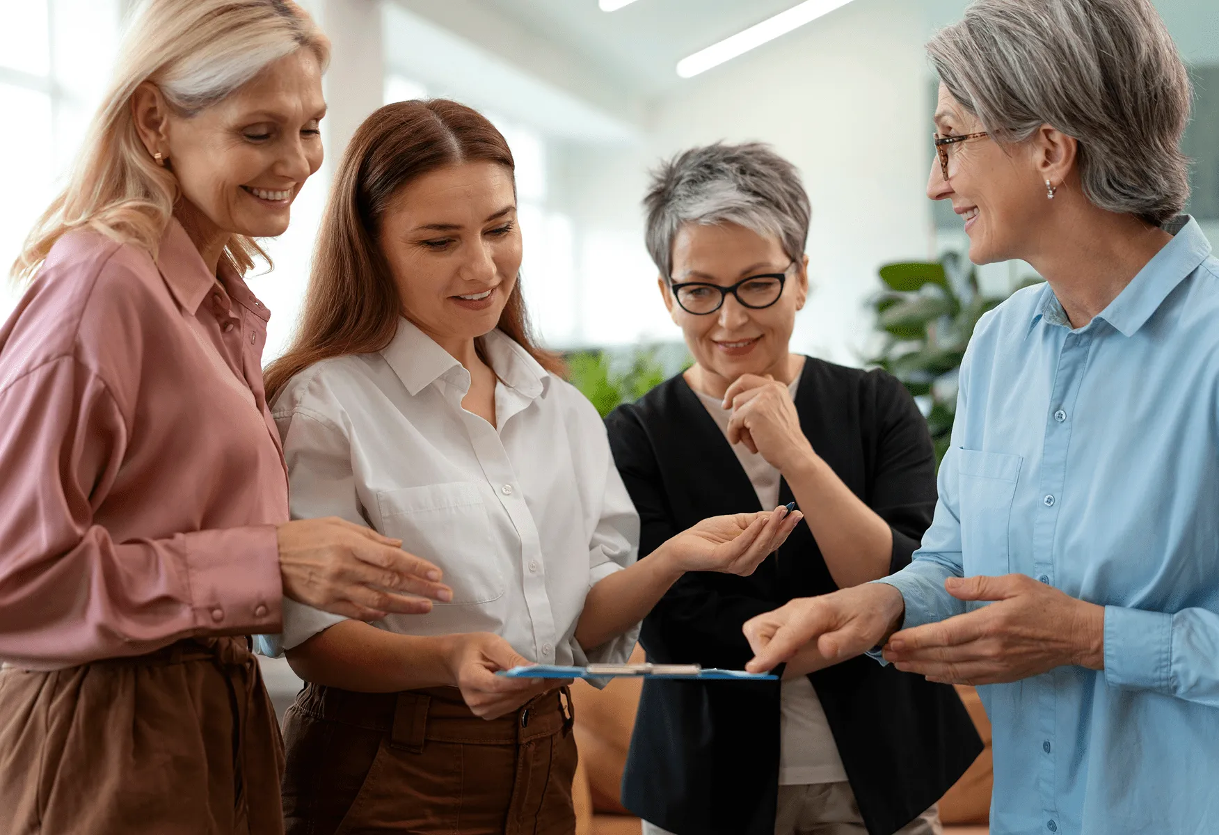 Četiri žene različitih generacija razgovaraju oko tableta u kancelariji sa biljkama u pozadini.
