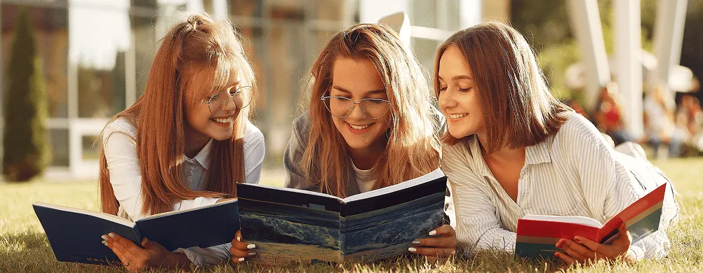 Tri devojke leže na travi i zajedno čitaju knjige na otvorenom tokom sunčanog dana.