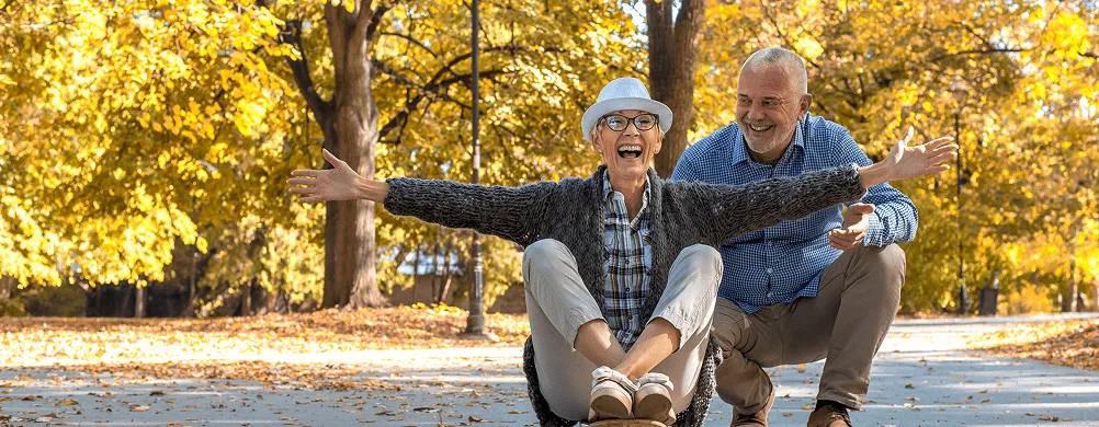 Srećan stariji par igra u parku sa zlatnim jesenjim lišćem, žena sedi na skejtu dok je muškarac gura.