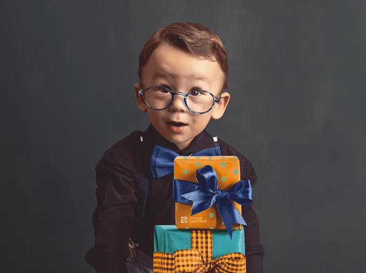 Mali dečak sa naočarima i plavom leptir mašnom drži dva poklon paketa sa plavim i narandžastim ukrasnim mašnama.