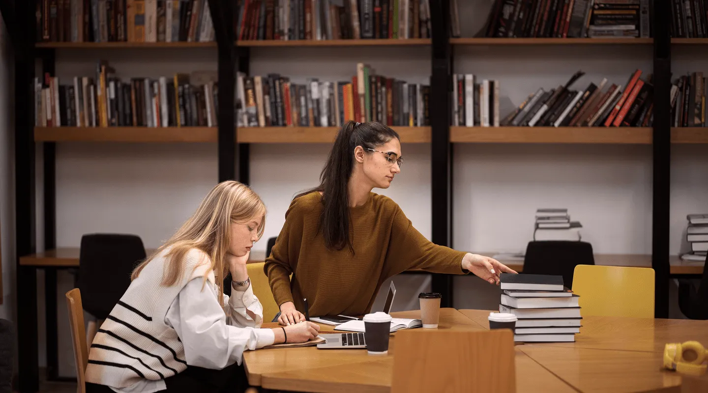 Dve žene koje uče za stolom u biblioteci, jedna piše dok druga pokazuje na gomilu knjiga.