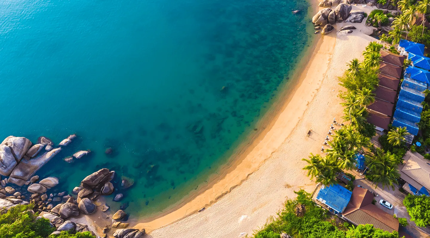 Pogled iz vazduha na tropsku plažu sa plavim krovovima, palmama, peskom i stjenovitim obalama kraj tirkiznog mora.