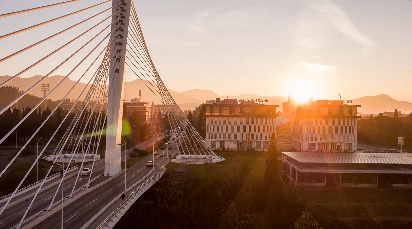 Sundown over a cable-stayed bridge with vehicles and nearby modern buildings with mountains in the background.