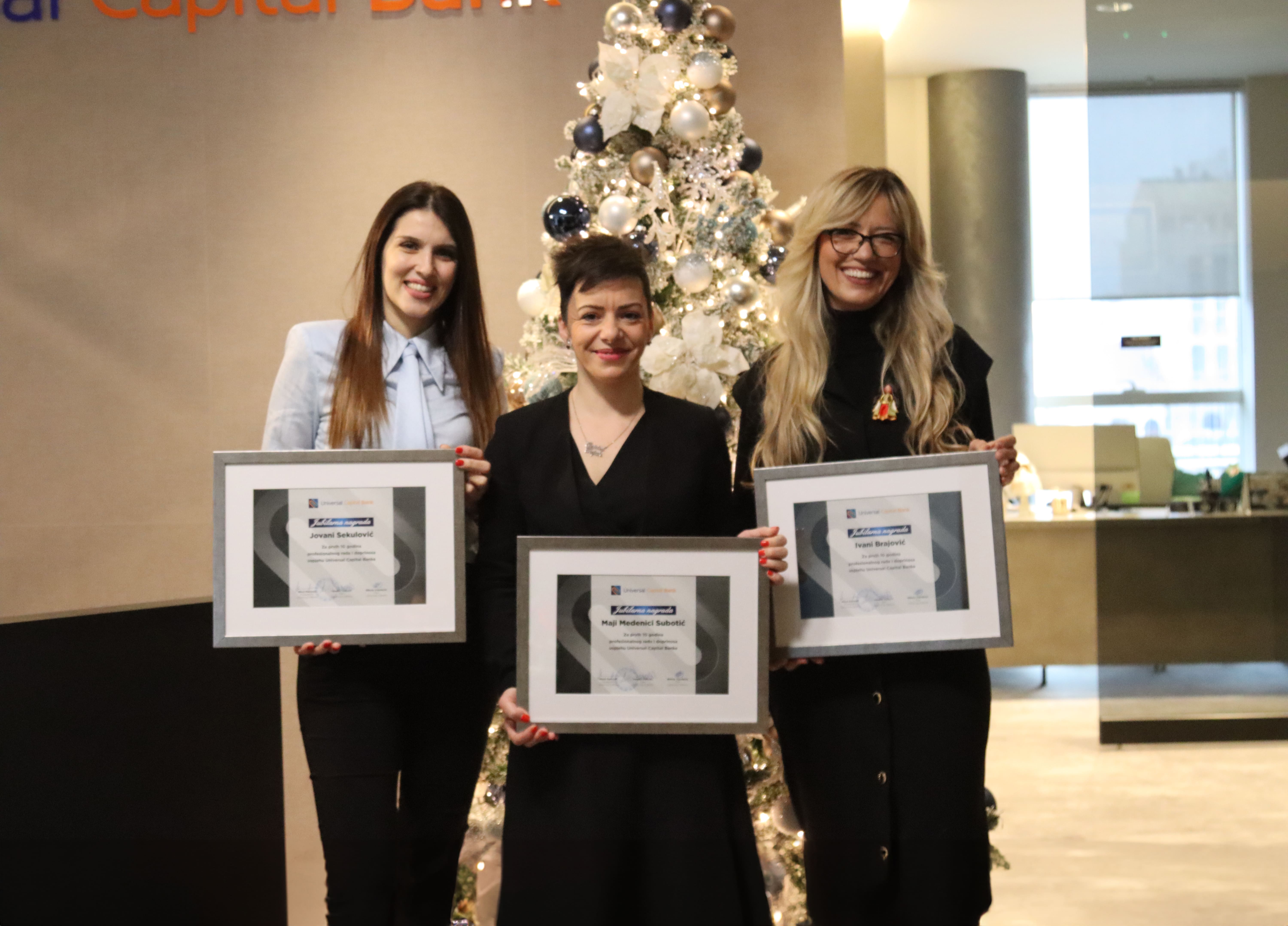 Tri žene stoje ispred ukrašene novogodišnje jelke u kancelariji, držeći okvirene sertifikate sa osmehom.