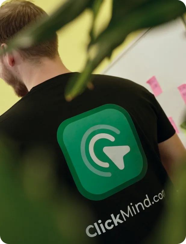 Man with short hair and beard wearing a black t-shirt with a green logo and the text 'ClickMind.com' on the back, standing indoors near a whiteboard with pink sticky notes.