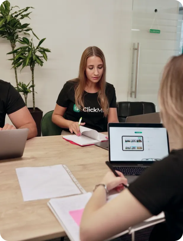 Three people working at a table in an office; one woman with long blonde hair in a black ClickMango shirt is writing in a notebook, another person is using a laptop, and a third person partially visible is also at the table.