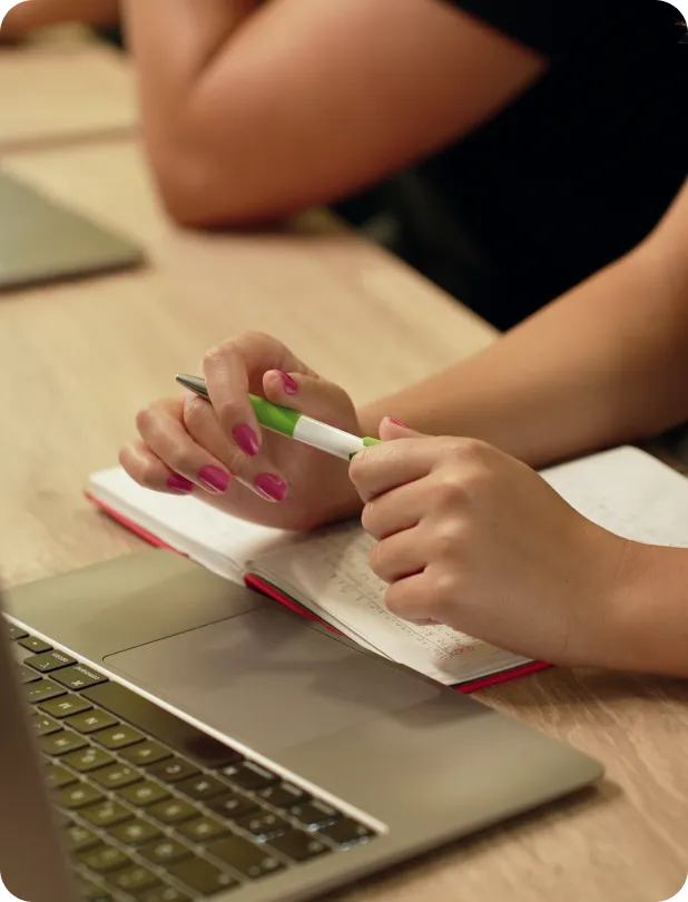Person with pink nail polish holding a green and white pen over an open notebook next to a laptop on a wooden table.