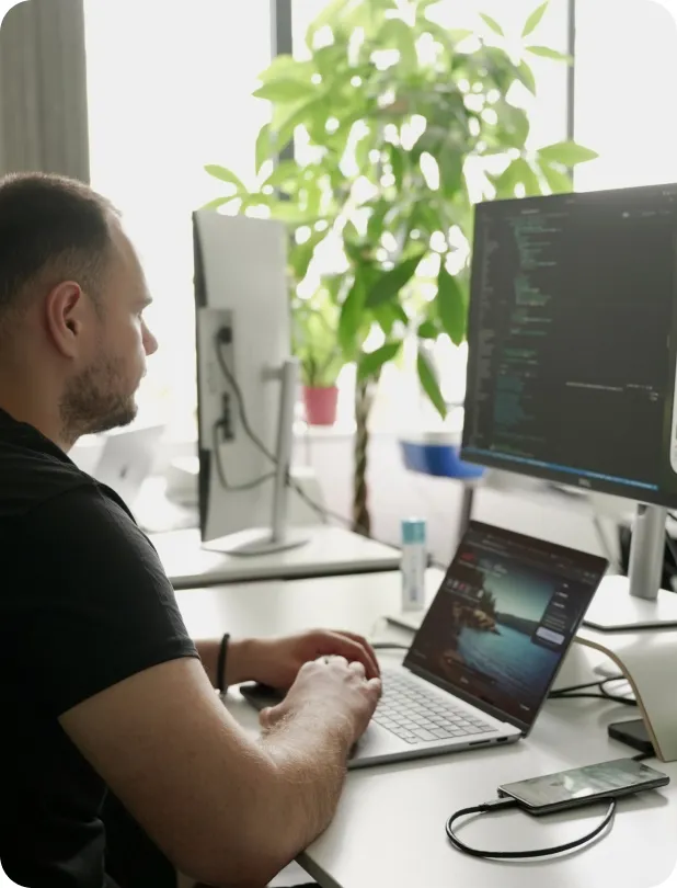Man in a black t-shirt typing on a laptop at a desk with a large monitor displaying code, a smartphone connected by a cable, and a green potted plant in the background near a bright window.