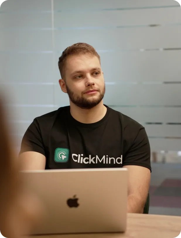 A young man with short hair and a beard wearing a black ClickMind t-shirt sits at a table with a silver Apple laptop in front of him, looking thoughtfully to the side.