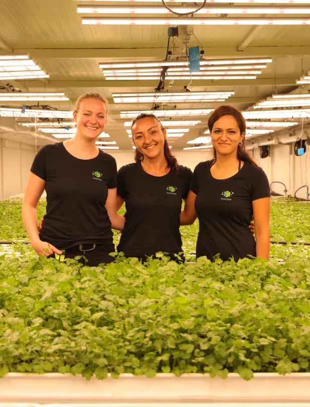 Three women standing closely together, smiling, in a large indoor hydroponic farm surrounded by green leafy plants under bright LED grow lights.