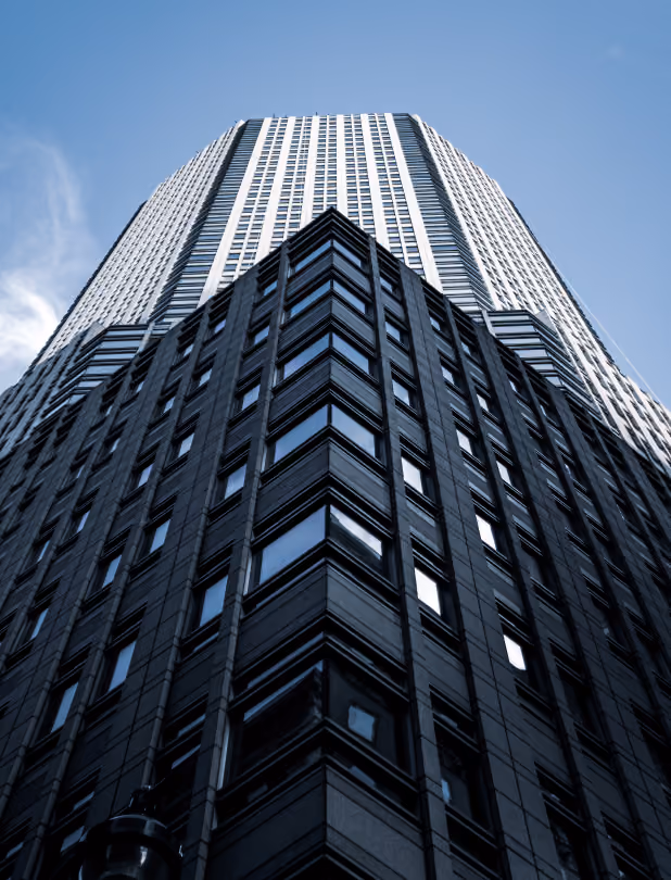 Low-angle view of a tall modern skyscraper with dark lower floors and lighter upper floors against a clear blue sky.