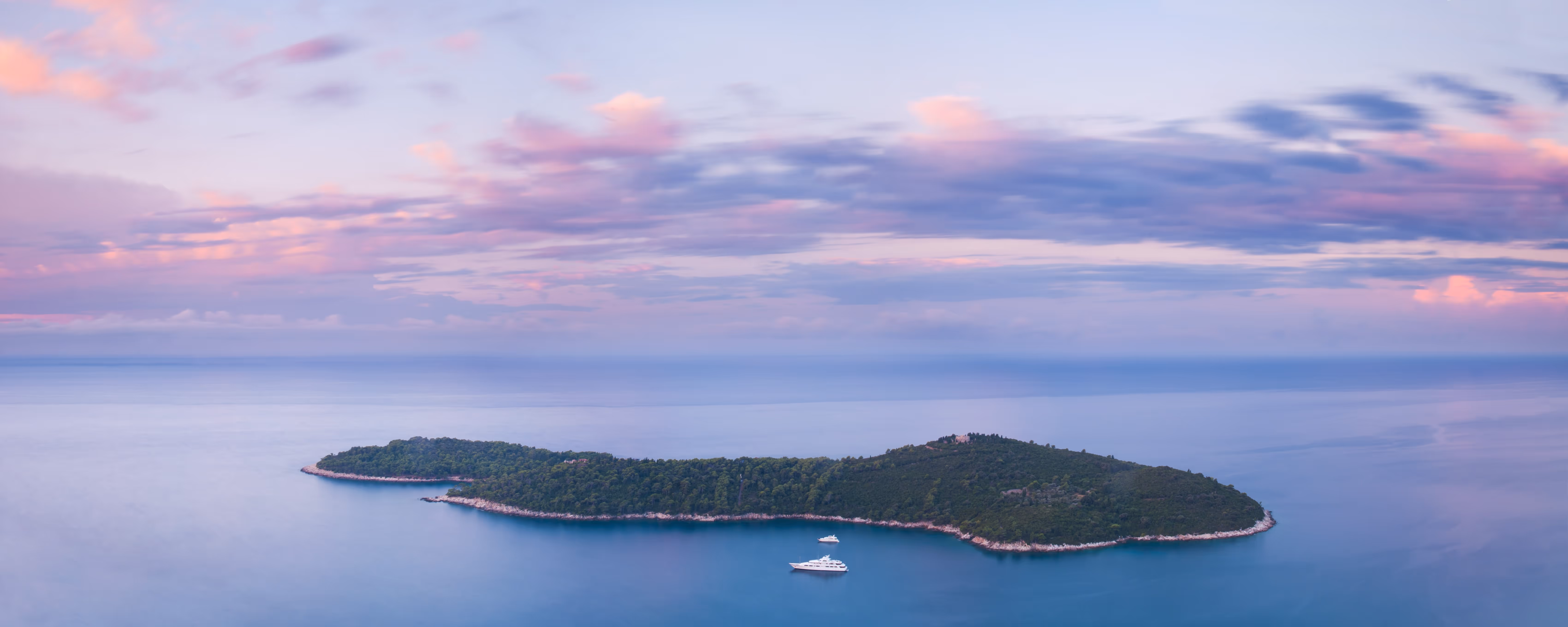 Aerial view of a small, forested island surrounded by calm water with two yachts anchored nearby under a pastel sunset sky.