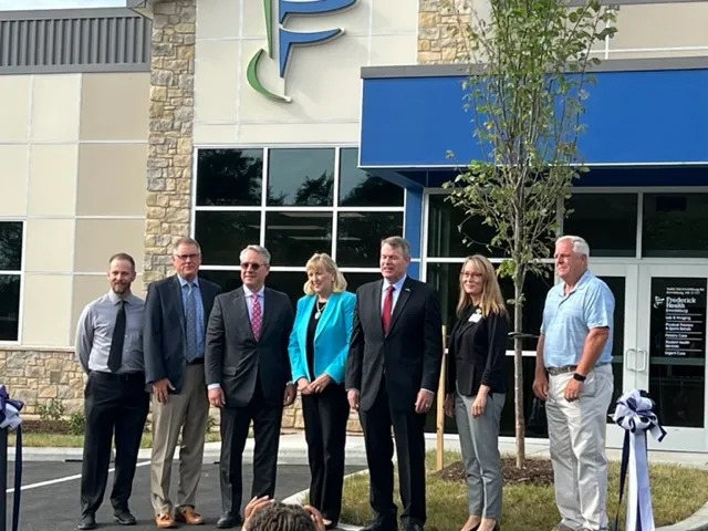 Seven people pose in front of a modern stone-and-glass building with a blue awning and 'F P' logo, flanked by ribbon bows and a small tree near the entrance