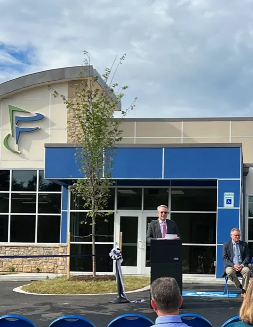 Outdoor event in front of blue-beige building with green-blue 'F' logo, featuring a man speaking at a podium and another seated, with blue chairs arranged for attendees