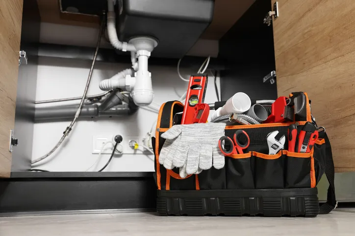 A plumber's tool bag filled with various tools, including a wrench, level, and PVC pipes, sitting on the floor in front of a cabinet with exposed plumbing