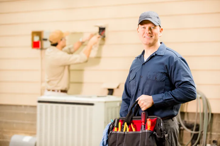 A smiling HVAC technician holds a tool bag in the foreground, with another technician working in the background