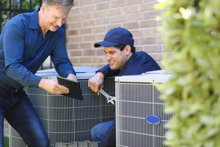 A smiling HVAC technician holding a tablet stands in front of a brick wall as a coworker works on an AC unit