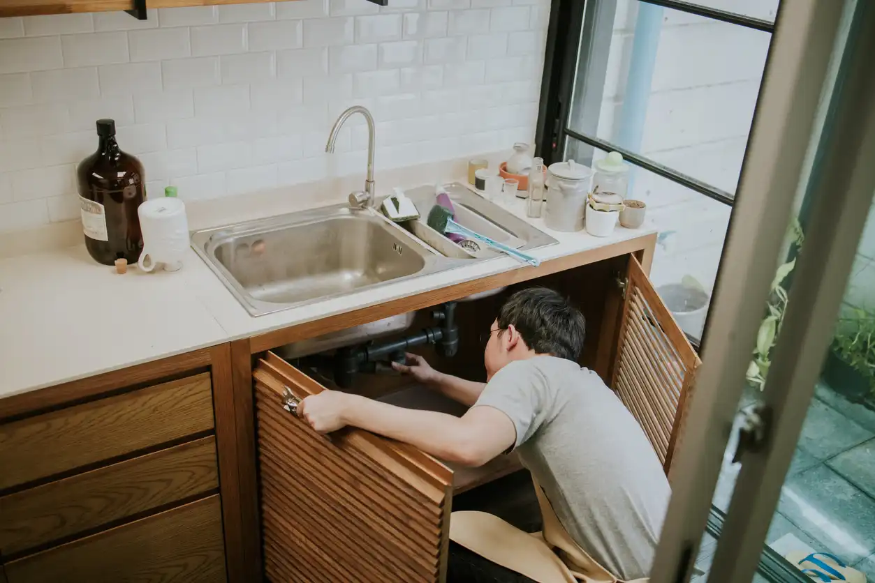 A plumber in a gray shirt and apron kneels in front of a modern kitchen sink, reaching into the open cabinet to work on the pipes.