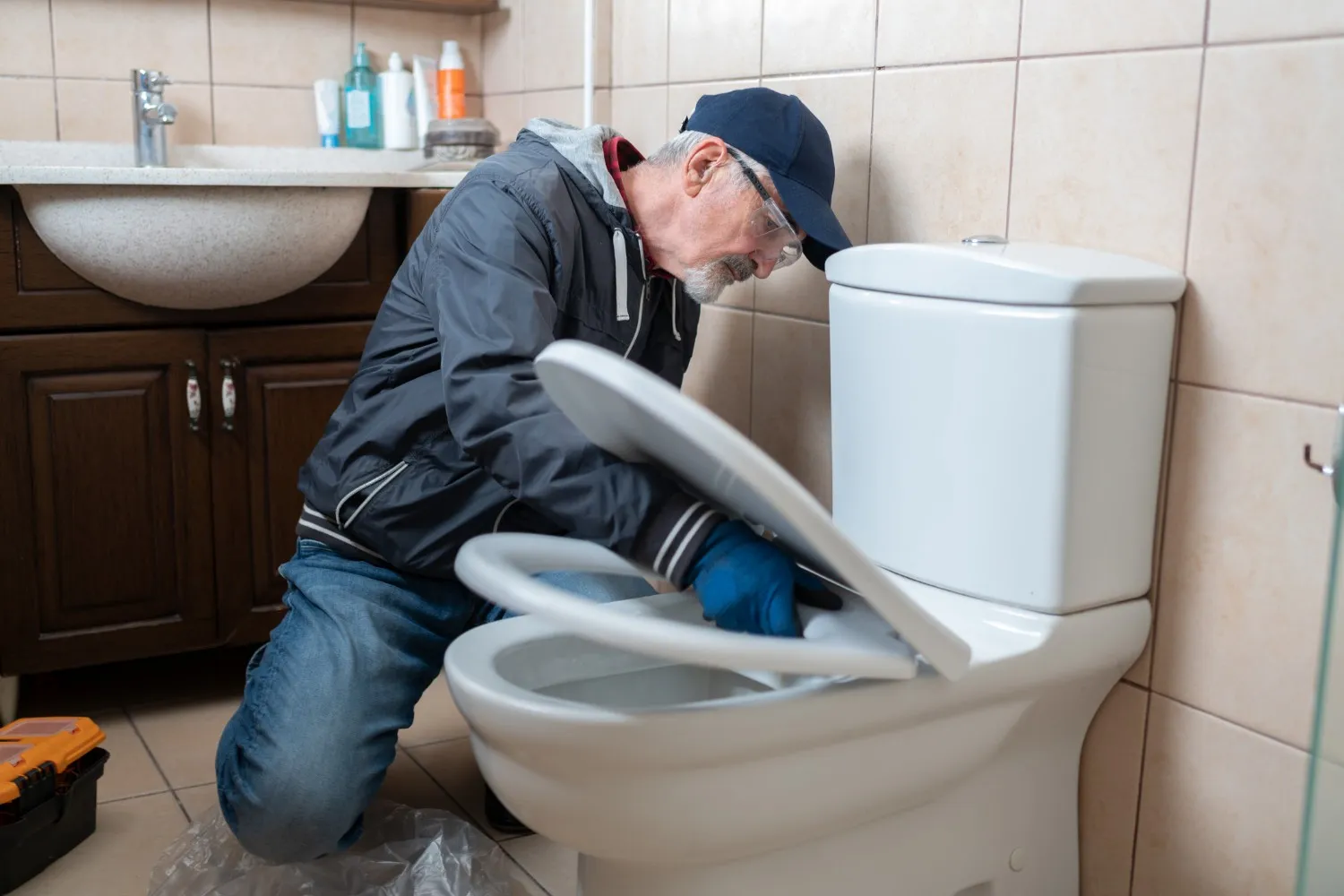 A senior male plumber in a black jacket and a baseball cap is kneeling and working on a toilet seat in a bathroom.