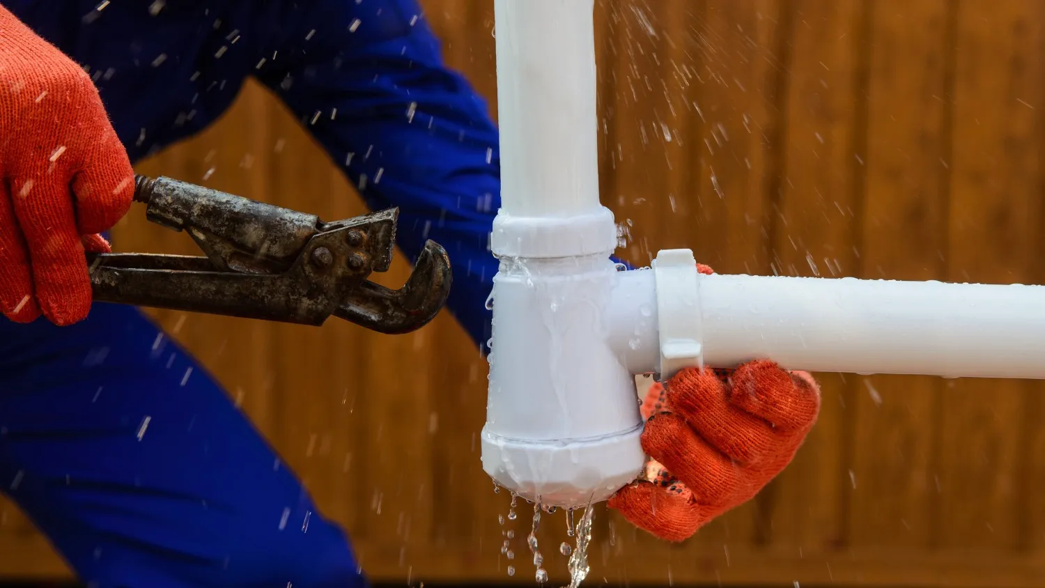 A close-up of a person wearing red gloves using a wrench to repair a leaking white PVC pipe, with water splashing out.