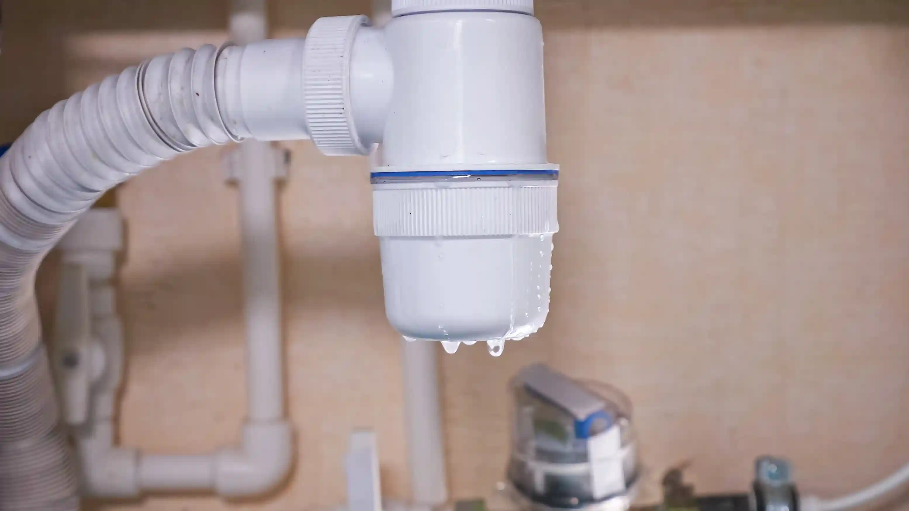 A close-up of a white plastic pipe under a sink with water dripping from the joint, indicating a leak