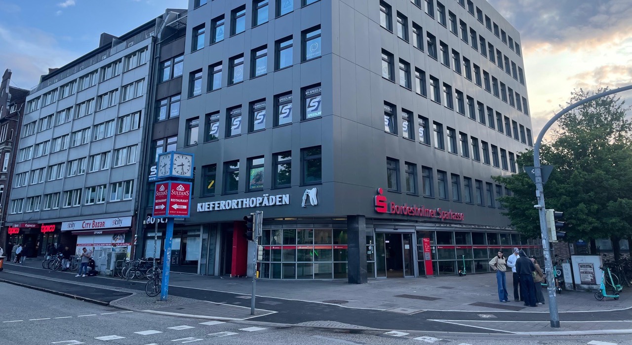 n einer modernen Zahnarztpraxis demonstriert eine Zahnmedizinische Fachkraft in schwarzem Polo-Shirt einer Patientin in weißer Bluse und blauer Hose die richtige Zahnpflege. Im Hintergrund ist eine türkisfarbene Wand und ein Bildschirm mit einer Zahndarstellung zu sehen. Beide lächeln während der Beratung.
